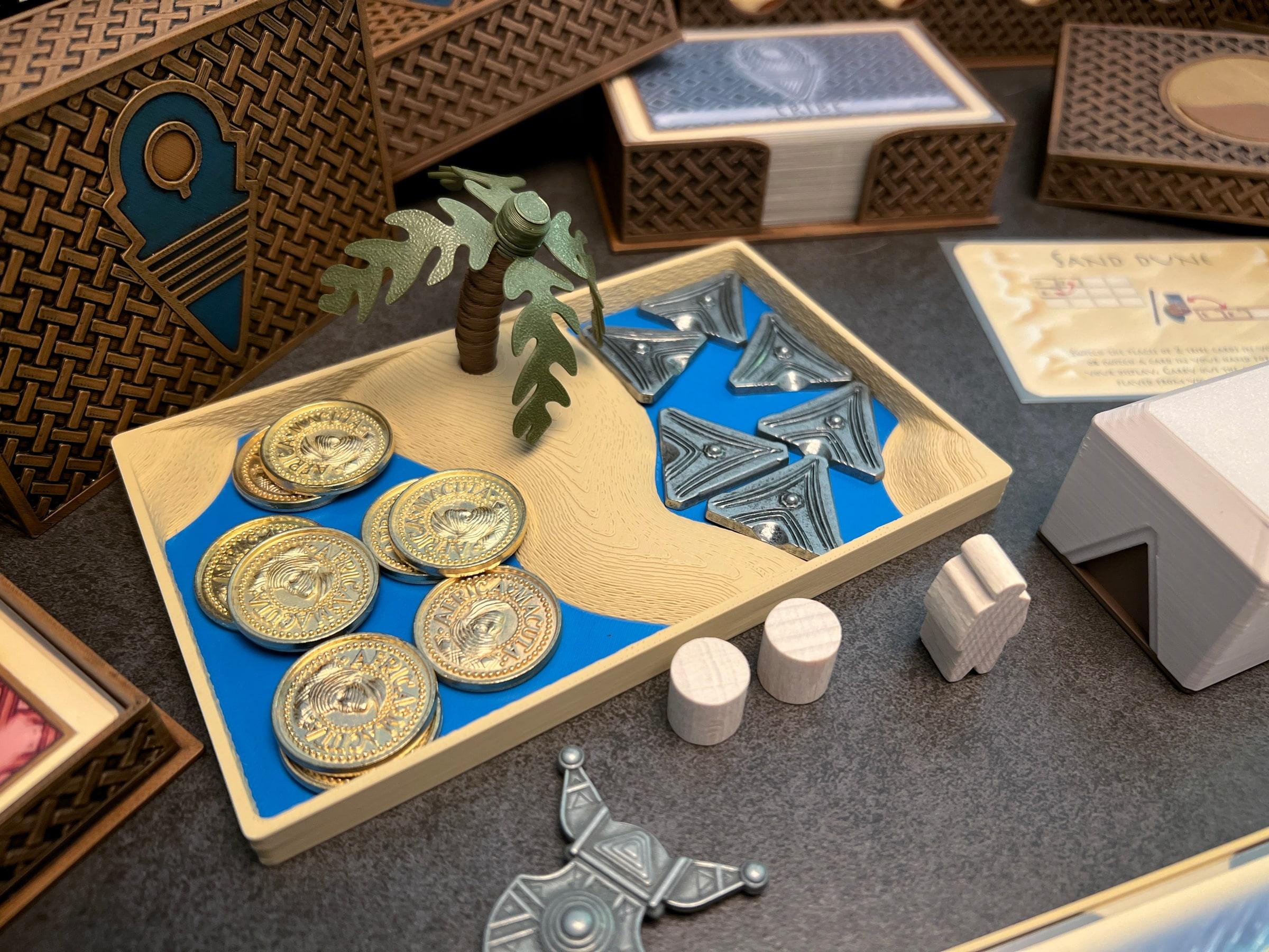 A 3D-printed Targi oasis tray: a sandy island with a palm tree, holding gold coins and silver tokens in blue water, among woven-pattern card boxes.